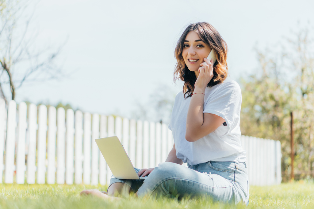 Laptop 'ın yanında akıllı telefondan konuşan ve çimlerin üzerinde oturan çekici serbest çalışanın seçici odağı - Fotoğraf, Görsel