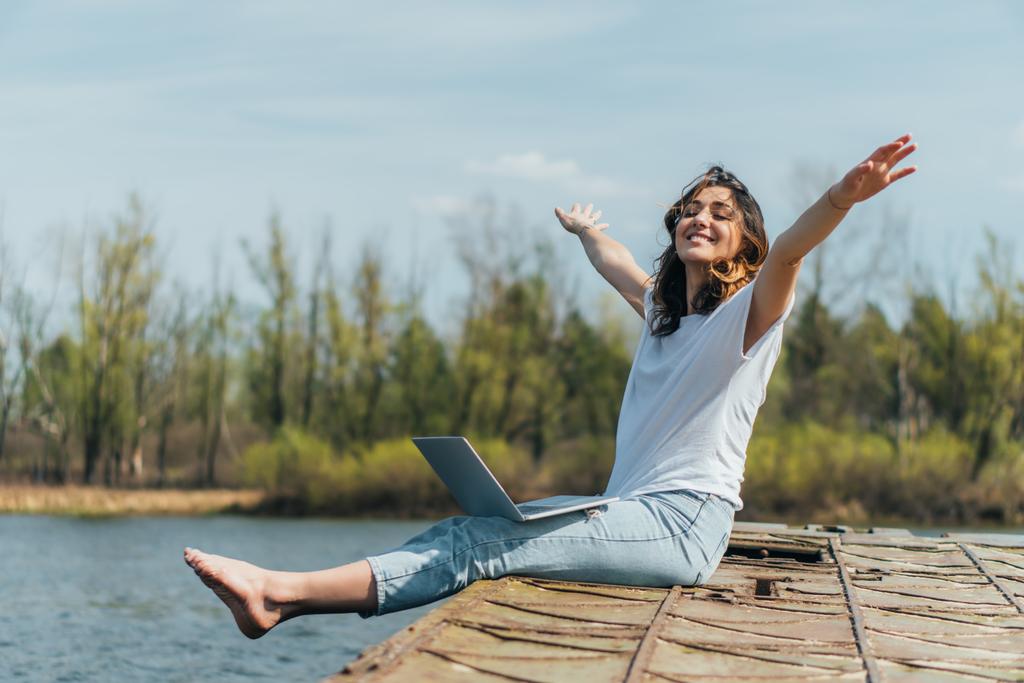 mulher alegre com as mãos estendidas sentado com laptop perto do lago
  - Foto, Imagem