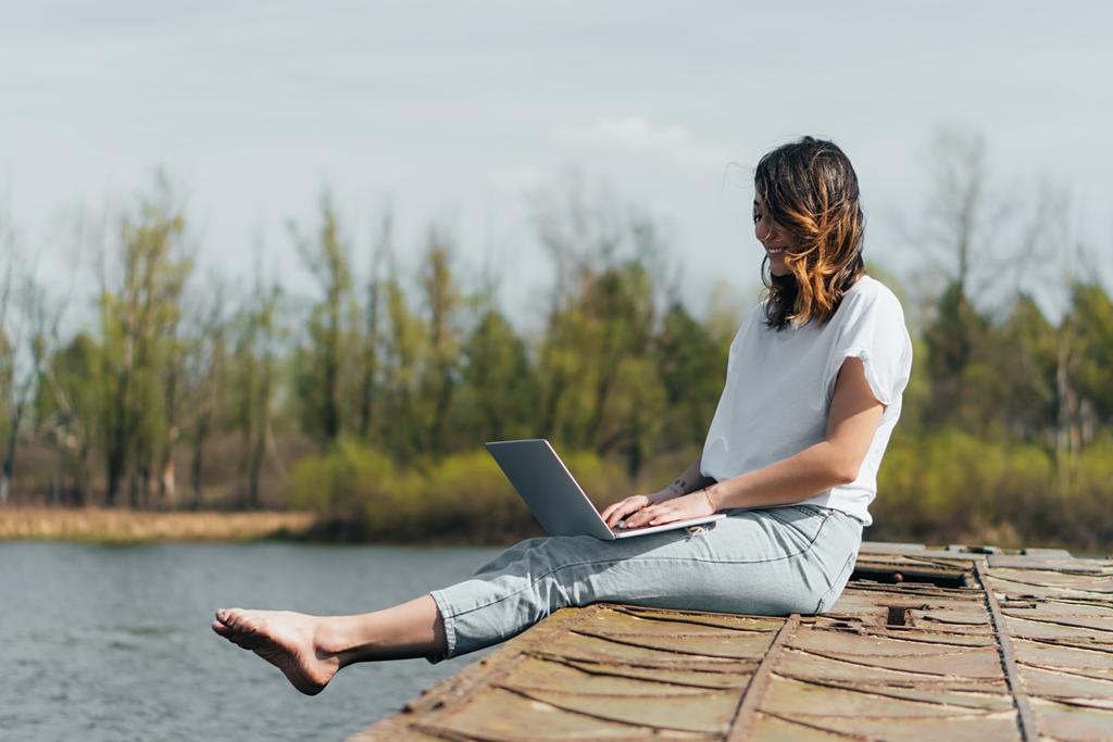 cheerful freelancer using laptop while networking near lake - Photo, Image