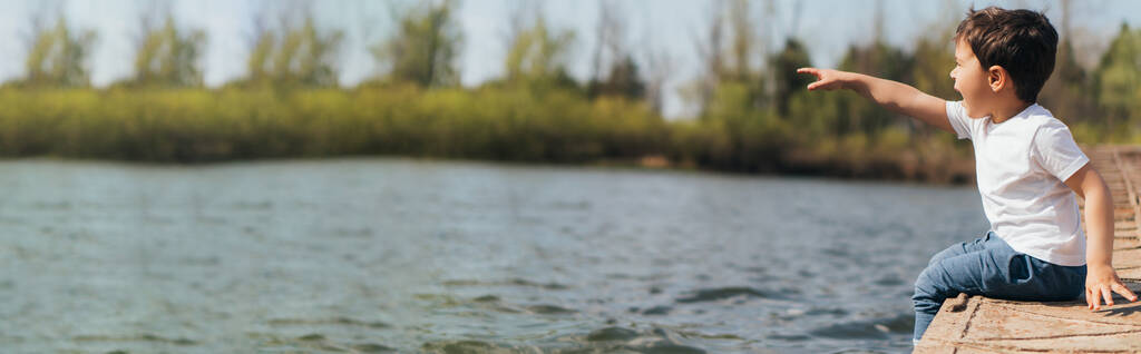 Website-Kopfzeile eines emotionalen Jungen, der mit dem Finger zeigt, während er in der Nähe des Flusses sitzt  - Foto, Bild