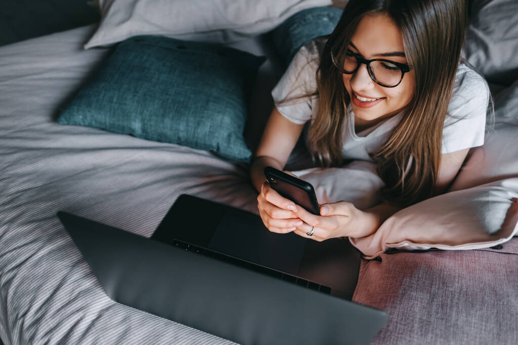 美しい若い笑顔の白人女性は自宅のベッドの上で休憩し、ノートパソコンやスマートフォンを使用しています。友達とのチャット、オンラインショッピング。高品質の写真 - 写真・画像