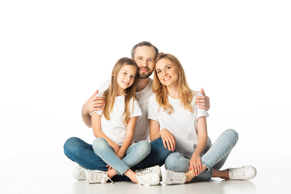 felice famiglia seduta sul pavimento con le gambe incrociate e abbracciando isolato su bianco - Foto, immagini