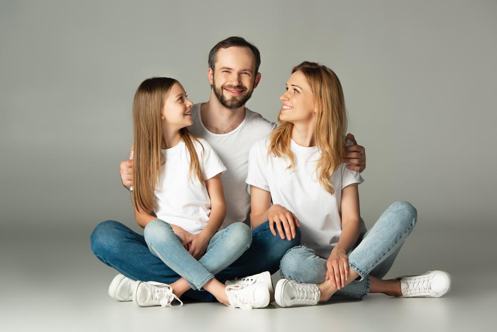 happy family sitting on floor with crossed legs and embracing on grey background - Photo, Image