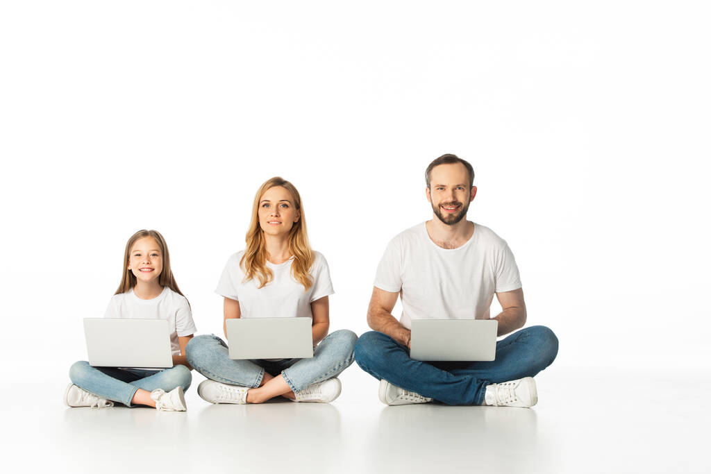 χαρούμενη οικογένεια κάθεται στο πάτωμα με φορητούς υπολογιστές σε σταυρωμένα πόδια που απομονώνονται σε λευκό - Φωτογραφία, εικόνα
