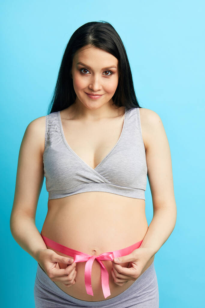 Красивая беременная женщина завязывая животик розовой лентой - Фото, изображение