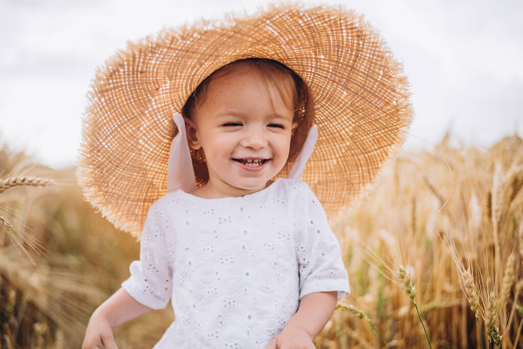 χαριτωμένο κοριτσάκι με όμορφο χαμόγελο που φοράει καλοκαιρινό ψάθινο καπέλο σε μια βόλτα στα χωράφια σιταριού. επιλεκτική εστίαση. οικογένεια, οι άνθρωποι έννοια - Φωτογραφία, εικόνα