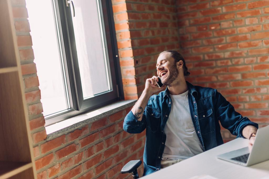 enfoque selectivo de hombre de negocios feliz hablando en el teléfono inteligente y el uso de ordenador portátil en la oficina
 - Foto, imagen