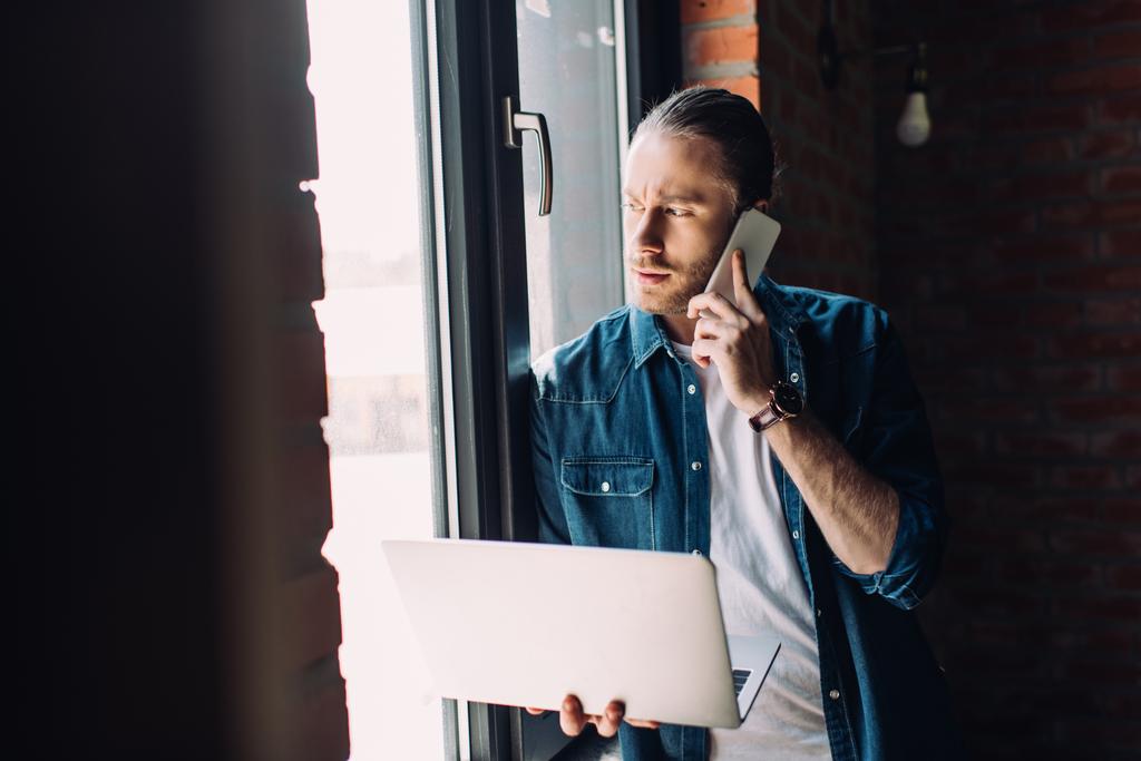 szelektív fókusz szakállas üzletember gazdaság laptop és beszél okostelefon ablak közelében  - Fotó, kép