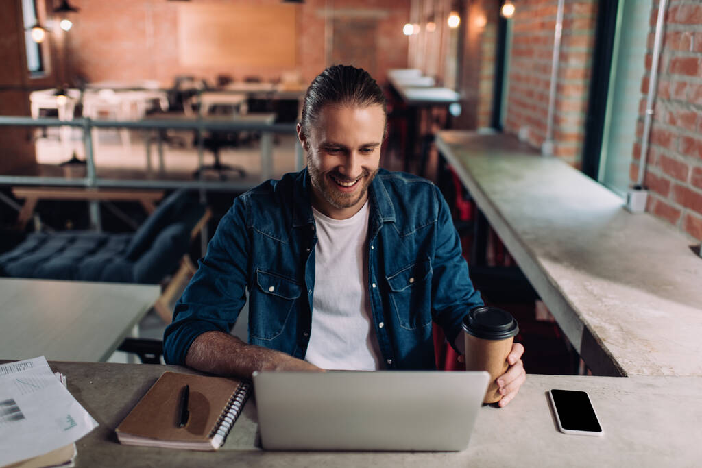 alegre hombre de negocios mirando a la computadora portátil y sosteniendo la taza de papel cerca del teléfono inteligente con pantalla en blanco
 - Foto, Imagen