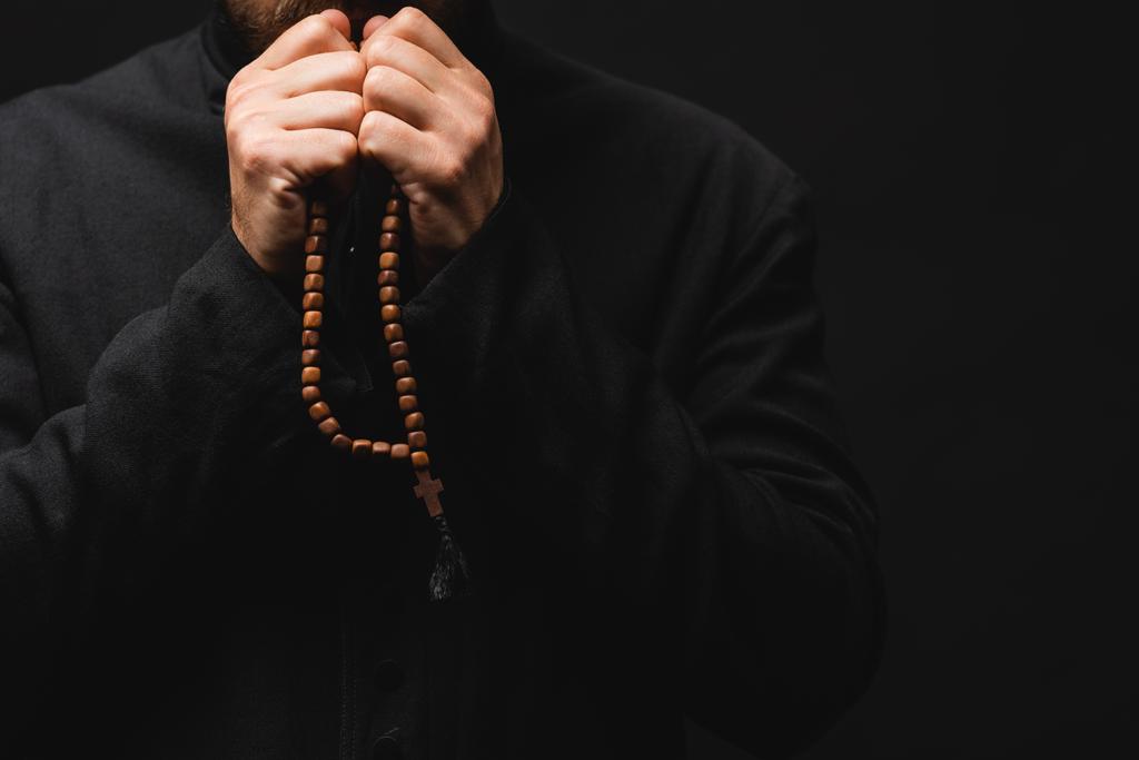 bijgesneden weergave van priester houden rozenkrans kralen in handen en bidden geïsoleerd op zwart  - Foto, afbeelding