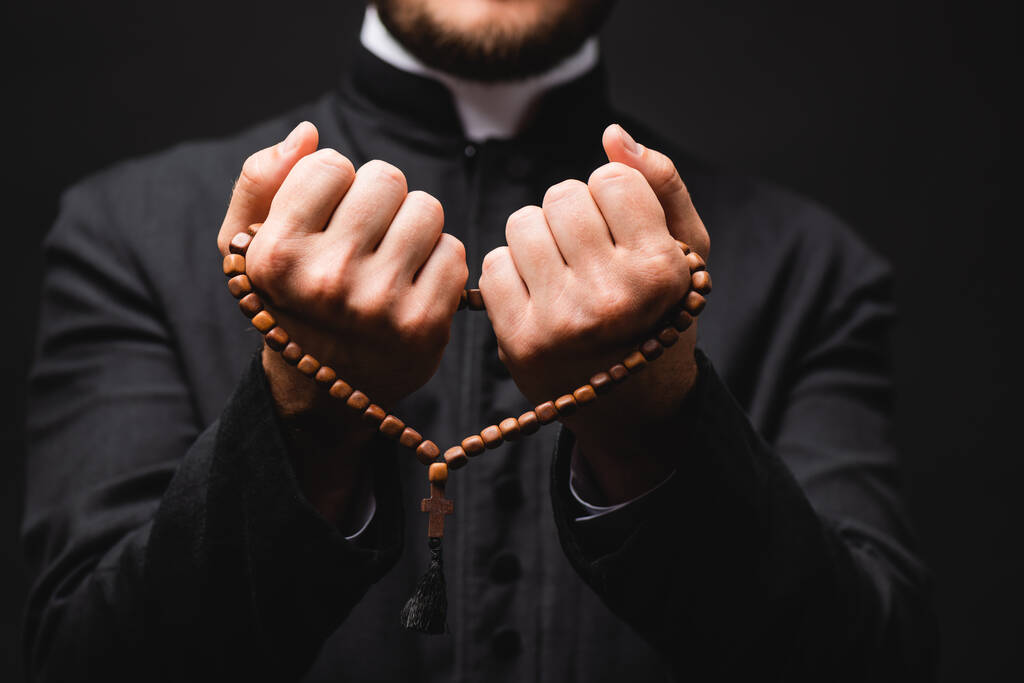 Selektiver Fokus des Pastors, der Rosenkranzperlen in den Händen hält und isoliert auf schwarz betet  - Foto, Bild