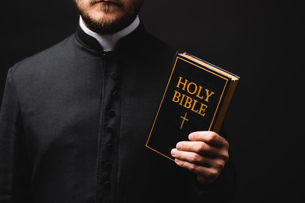 abgeschnittene Ansicht eines Priesters mit heiliger Bibel isoliert auf schwarz  - Foto, Bild