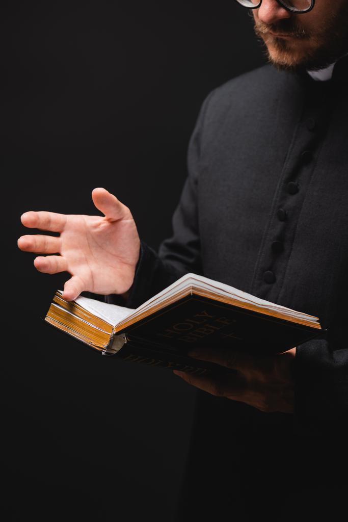 καλλιεργημένη άποψη του ιερέα που κρατά την Αγία Γραφή και gesturing απομονώνονται σε μαύρο  - Φωτογραφία, εικόνα