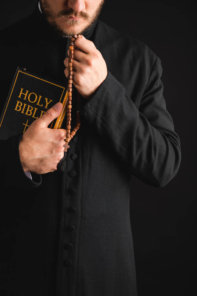 vista recortada del sacerdote barbudo sosteniendo la sagrada Biblia y rosario perlas en las manos aisladas en negro
  - Foto, imagen
