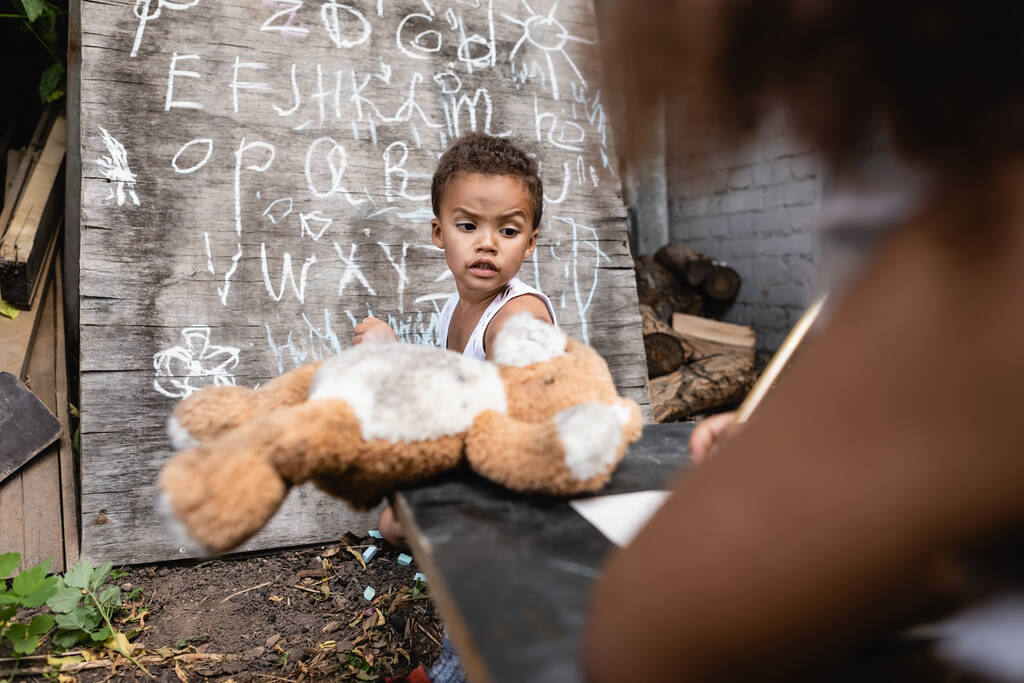 valikoiva painopiste afrikkalainen amerikkalainen poika tilalla pehmeä lelu lähellä lapsi ja liitutaulu  - Valokuva, kuva