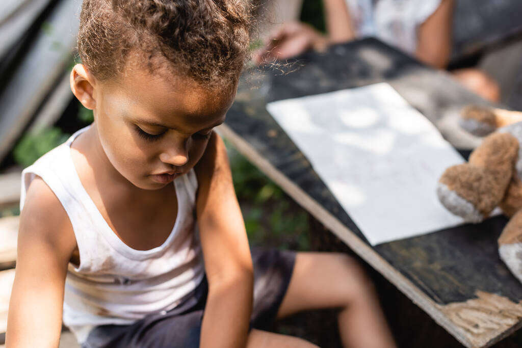 selective focus of poor african american boy looking down near table with paper  - Photo, Image