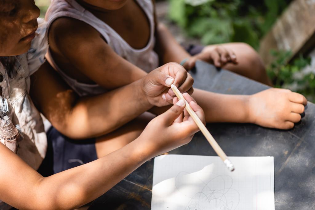 обрізаний вид бідної афроамериканської дитини, що тримає олівець біля блокнота на столі і дитини
 - Фото, зображення