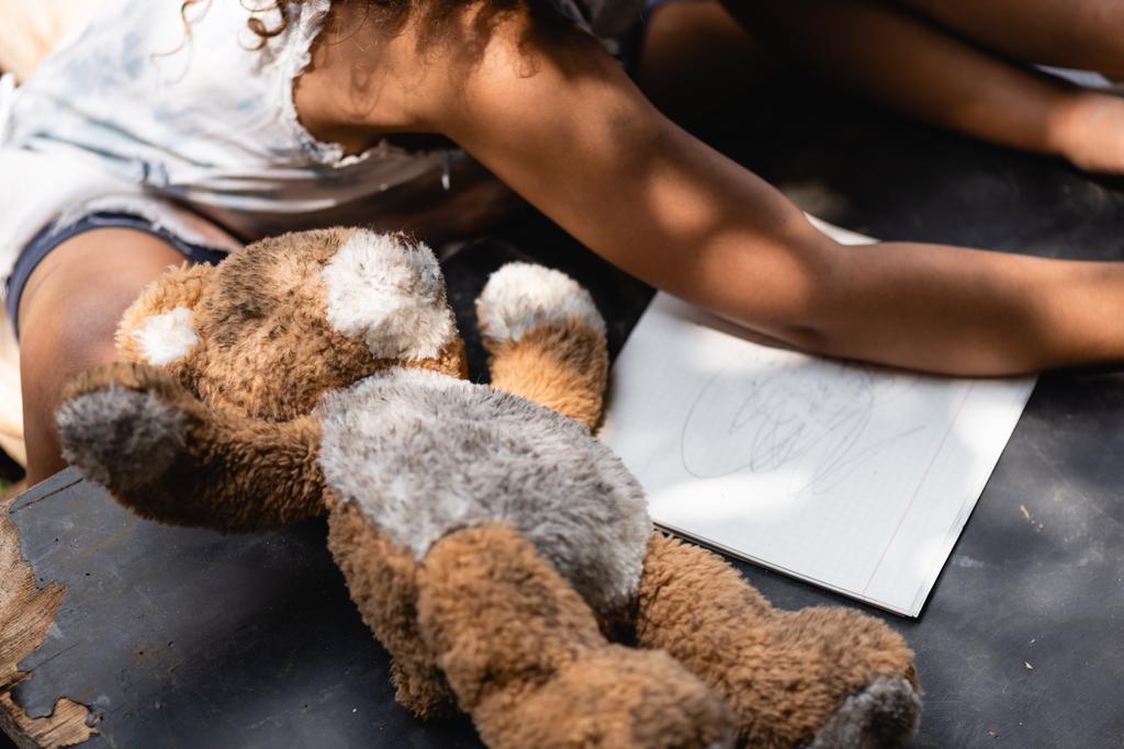 обрізаний вид на бідну афроамериканську дитину біля плюшевого ведмедя і блокнота на столі
  - Фото, зображення
