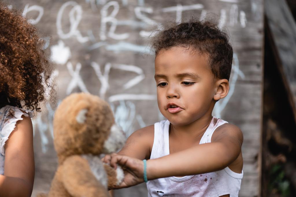 foco seletivo do pobre menino afro-americano brincando com urso de pelúcia sujo perto da criança
 - Foto, Imagem