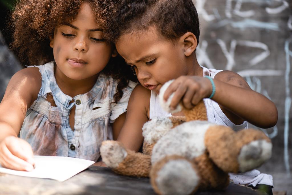 アフリカ系アメリカ人の子供が汚いテディベアで遊んでいる弟の近くで書いている間に  - 写真・画像