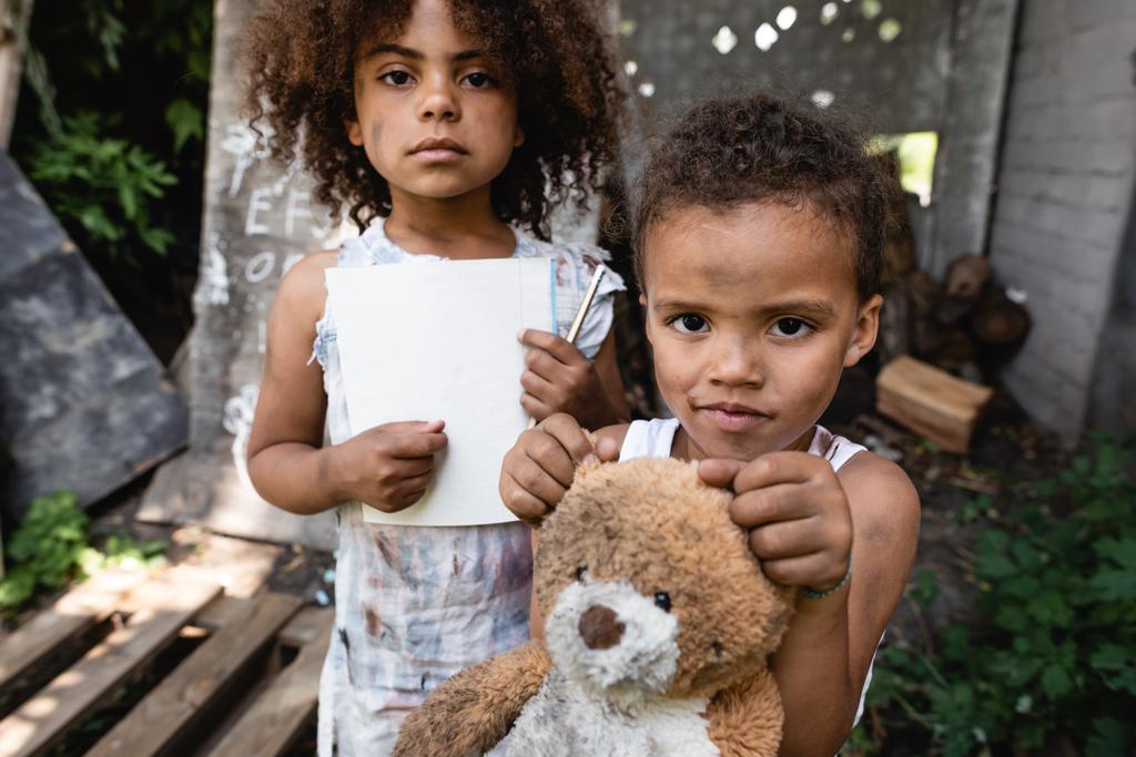 selectieve focus van arme Afro-Amerikaanse kind met blanco papier en potlood in de buurt van broer met teddybeer  - Foto, afbeelding