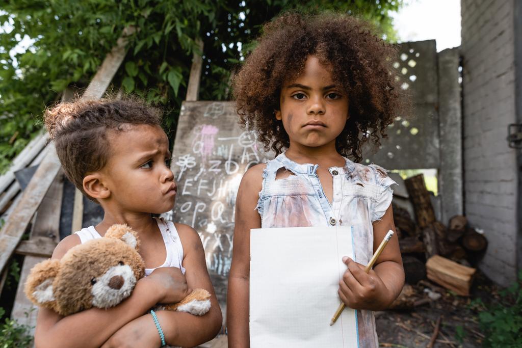 szomorú és szegény afro-amerikai gyerek kezében üres papír és ceruza közelében testvére játékmackó  - Fotó, kép