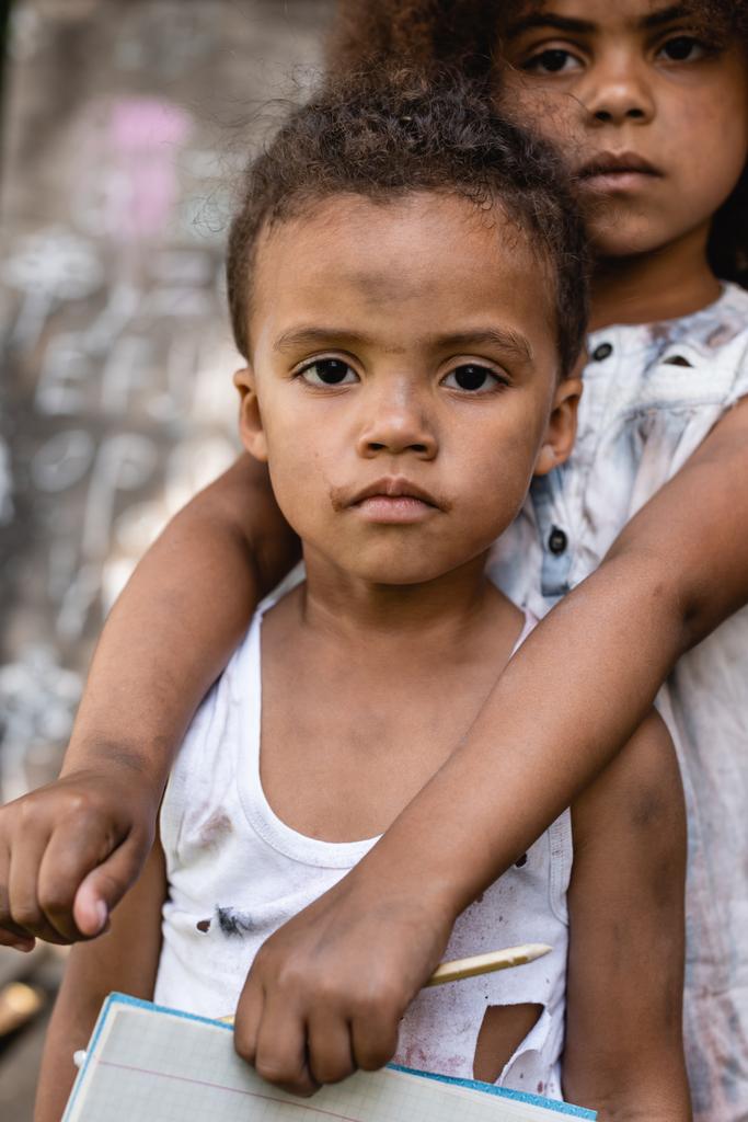 trauriges afrikanisch-amerikanisches Kind hält Bleistift und umarmt armen Bruder in zerrissenen Kleidern  - Foto, Bild