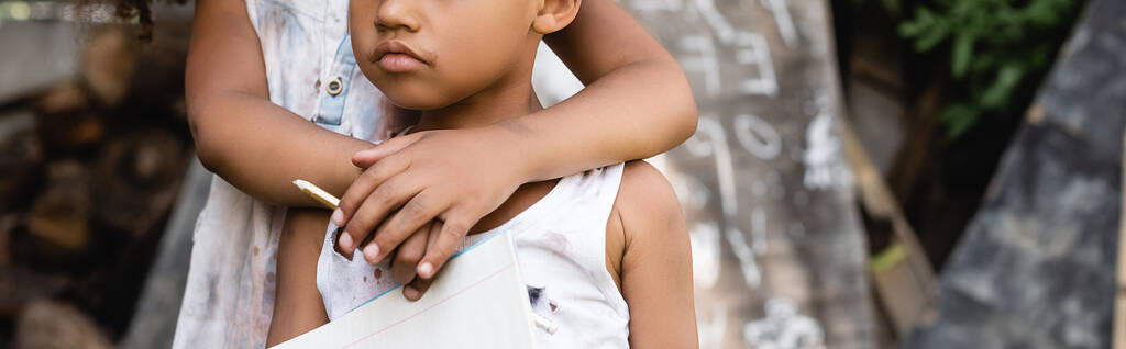 Panoramiczny plon afrykańskiego dziecka trzymającego ołówek i przytulającego biednego brata w podartych ubraniach  - Zdjęcie, obraz