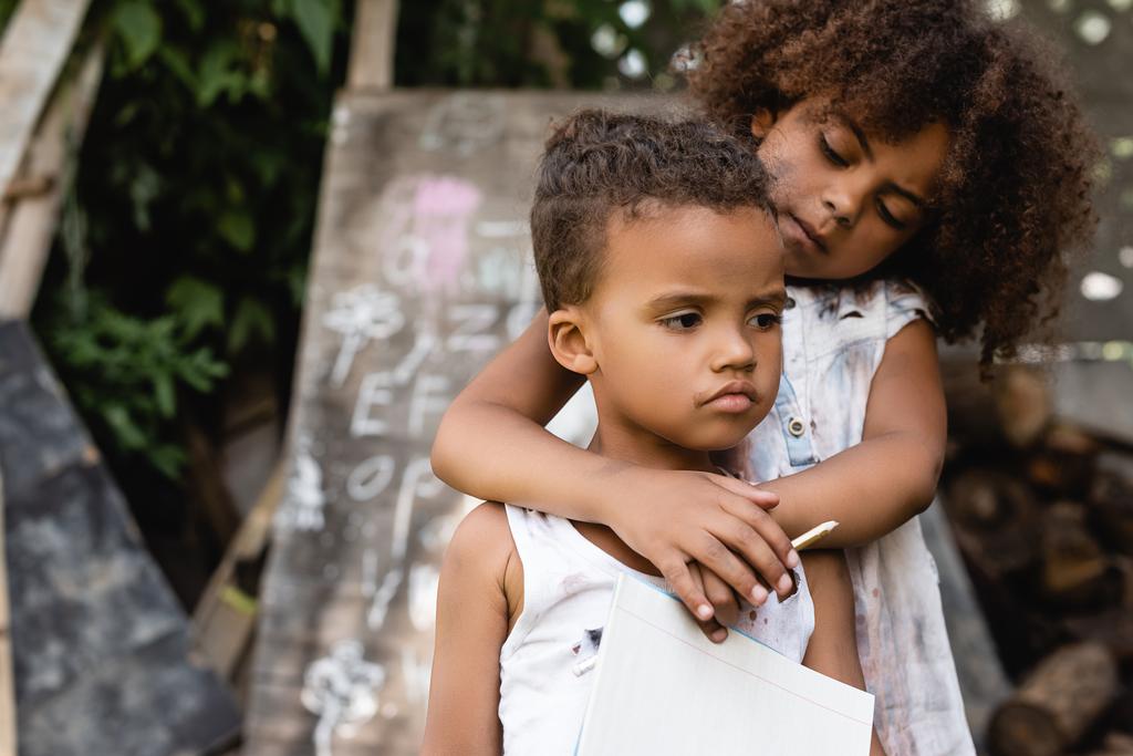 armes afrikanisch-amerikanisches Kind hält Bleistift in der Hand und umarmt traurigen Bruder in zerrissenen Kleidern  - Foto, Bild