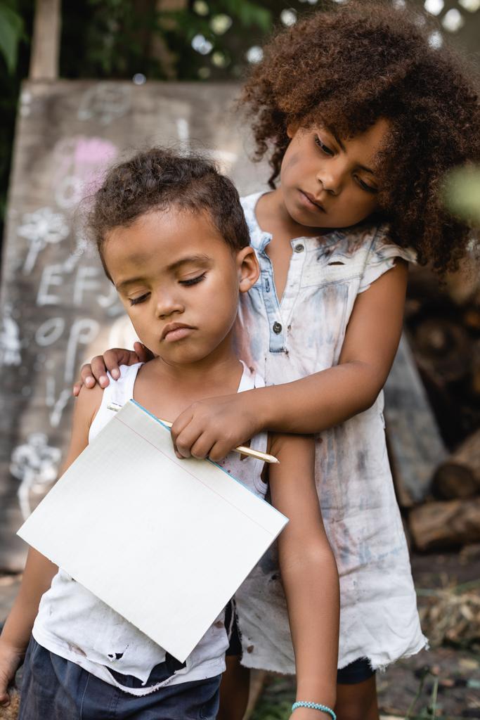 pobre afroamericano niño sosteniendo en blanco cuaderno y lápiz mientras toca triste hermano en rasgado ropa
  - Foto, imagen