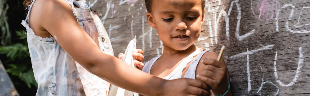 Kara tahtanın yanında dikilen ve ellerinde kalem tutan yırtık elbiseli fakir Afrikalı Amerikalı çocukların panoramik ürünleri.  - Fotoğraf, Görsel