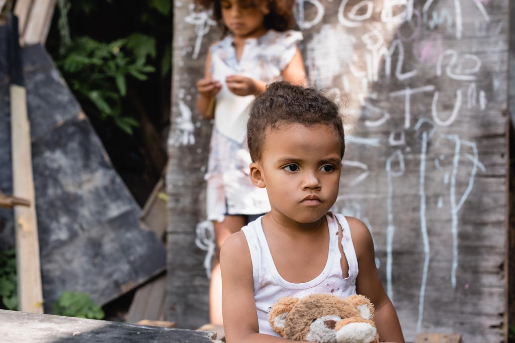 valikoiva painopiste köyhien afrikkalainen amerikkalainen lapsi revitty vaatteita istuu pehmeä lelu lähellä liitutaulu ja sisko - Valokuva, kuva