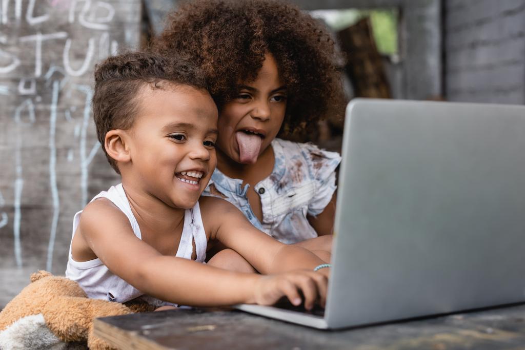 selektywne skupienie biednego afrykańskiego dziecka wystającego z języka w pobliżu szczęśliwego dziecka korzystającego z laptopa na zewnątrz  - Zdjęcie, obraz