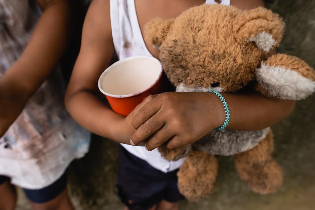 Gehakt uitzicht op vuil Afrikaans-Amerikaanse kinderen met teddybeer en beker bedelen aalmoezen op stedelijke straat  - Foto, afbeelding