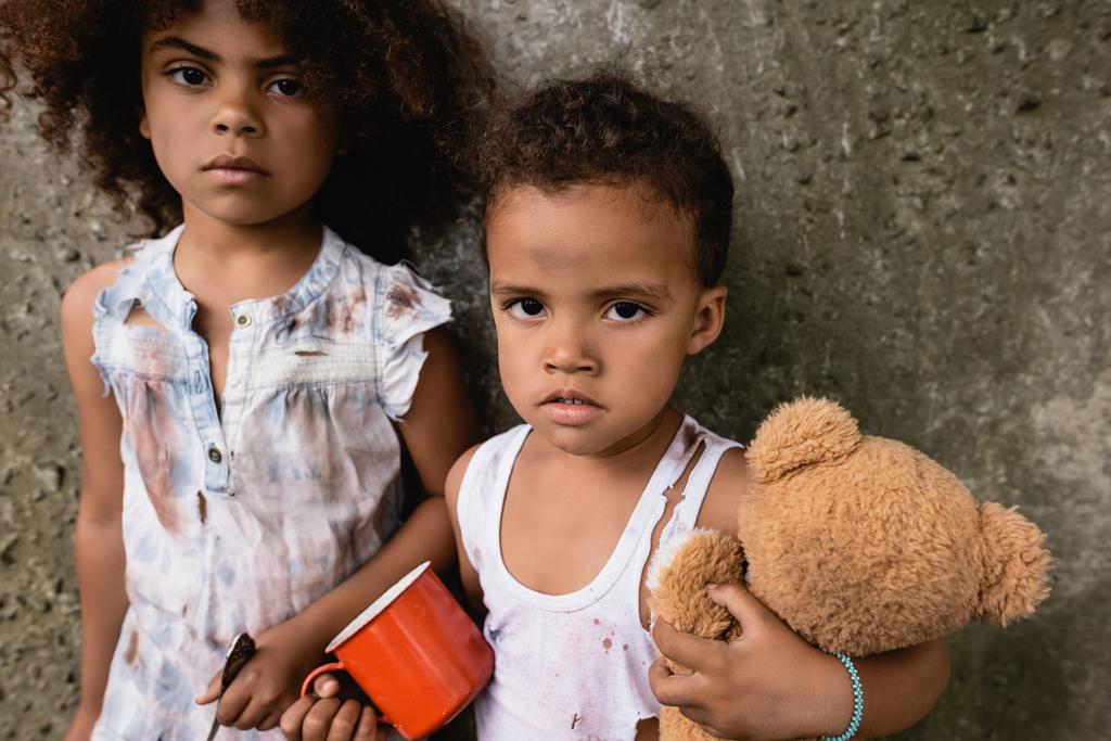 Καημένα παιδιά από την Αφρική σε βρώμικα ρούχα με μεταλλικό κύπελλο και αρκουδάκι κοιτάζοντας την κάμερα κοντά σε τσιμεντένιο τοίχο σε εξωτερικούς χώρους  - Φωτογραφία, εικόνα