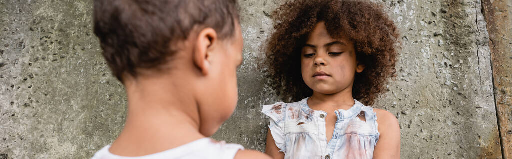 Панорамный урожай африканского американского ребенка в грязной одежде, стоящего возле брата и бетонной стены на городской улице  - Фото, изображение
