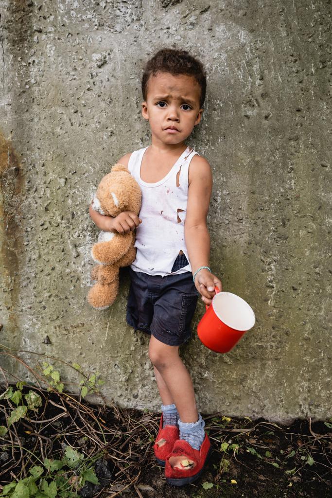 Δυσαρεστημένο αφροαμερικανό παιδί με βρώμικα ρούχα ζητιανεύει ελεημοσύνη κοντά σε τσιμεντένιο τοίχο στην παραγκούπολη.  - Φωτογραφία, εικόνα