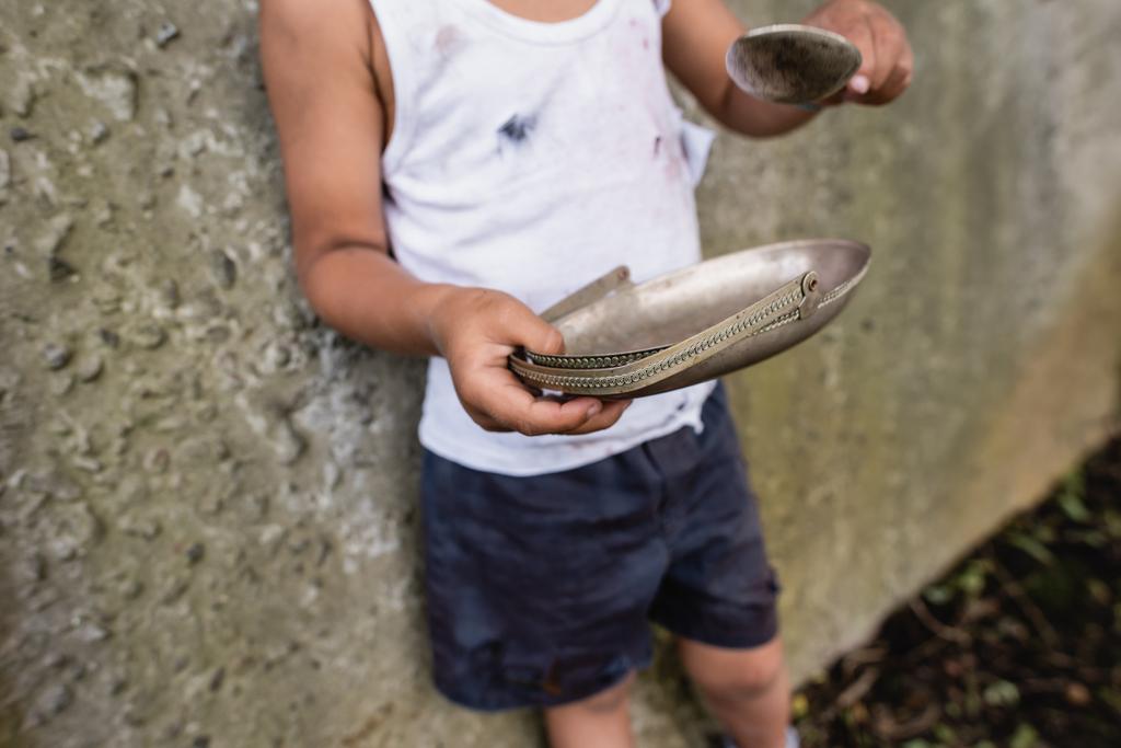 Cropped view of poor african american kid holding dirty spoon and plate on urban street  - Photo, Image
