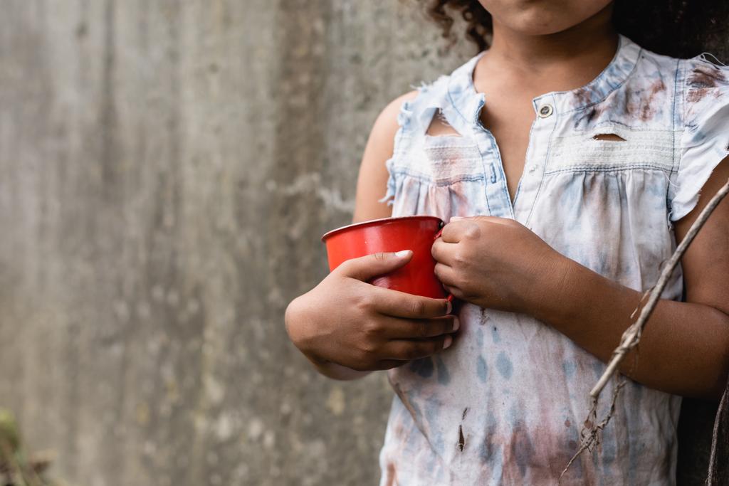 Обрізаний вид бідної афроамериканської дитини в брудному одязі, що тримає металеву чашку на міській вулиці
  - Фото, зображення