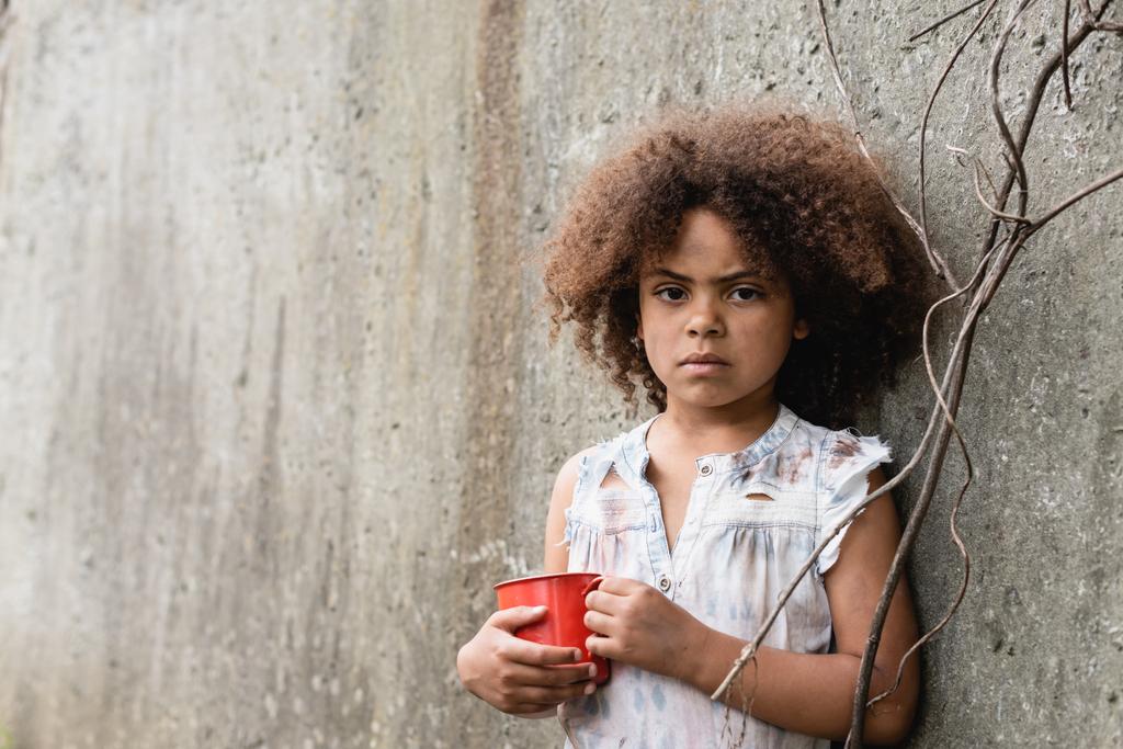 Καημένο αφροαμερικανό παιδί που κοιτάζει την κάμερα κρατώντας ένα μεταλλικό κύπελλο κοντά σε τσιμεντένιο τοίχο στην παραγκούπολη  - Φωτογραφία, εικόνα