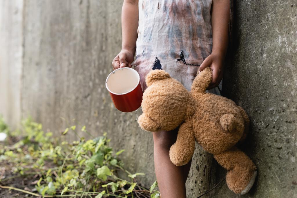 Cropped view of beggar african american child holding teddy bear and cup near concrete wall on urban street  - Photo, Image