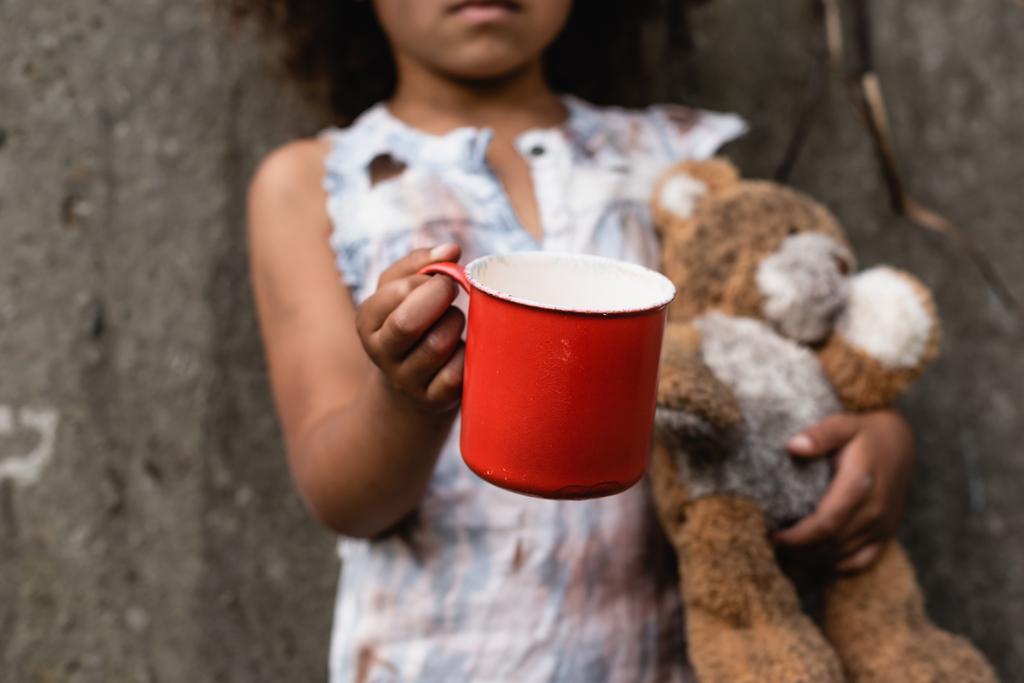 Vista recortada de un pobre niño afroamericano sosteniendo una taza de metal y un oso de peluche mientras mendigaba limosna en la calle urbana
  - Foto, Imagen