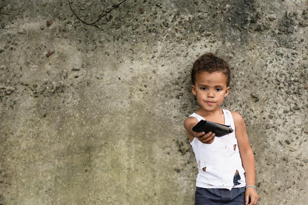 都市部のコンクリート壁の近くに財布を示す貧しいアフリカ系アメリカ人の少年の選択的な焦点  - 写真・画像