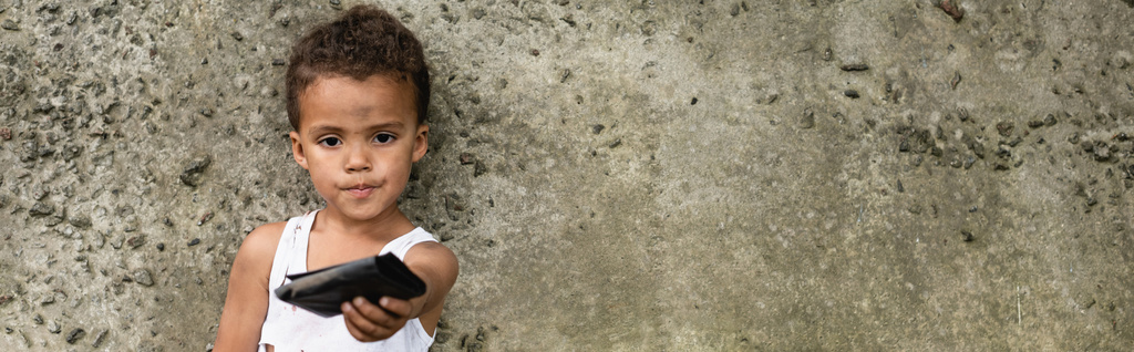 Πανοραμική φωτογραφία αφροαμερικανού παιδιού που κρατάει πορτοφόλι κοντά σε τσιμεντένιο τοίχο σε αστικό δρόμο  - Φωτογραφία, εικόνα