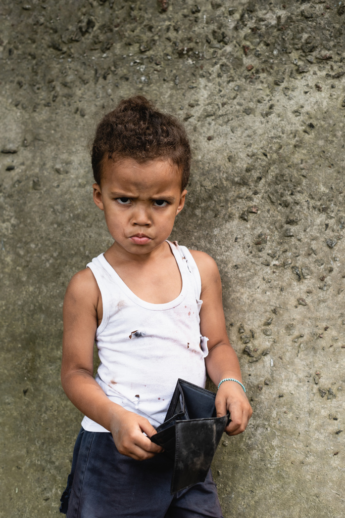 Θυμωμένο αφροαμερικανό παιδί με βρώμικα ρούχα που κρατάει άδειο πορτοφόλι στον αστικό δρόμο  - Φωτογραφία, εικόνα