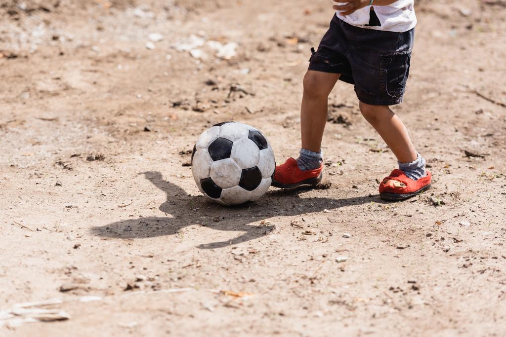 Vista recortada del pobre chico afroamericano jugando con pelota de fútbol en camino sucio en la calle urbana
  - Foto, Imagen