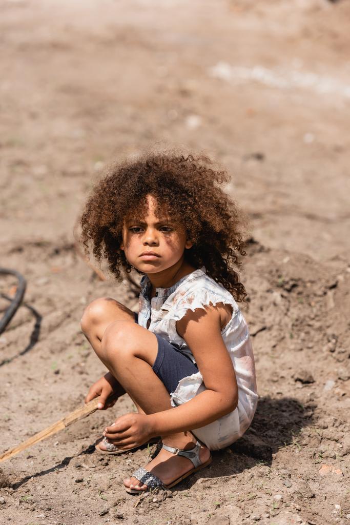 Pobre criança afro-americana segurando galho de madeira enquanto brincava na estrada suja na rua urbana
  - Foto, Imagem