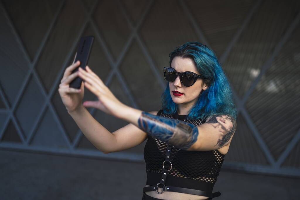 Eine Europäerin mit Tätowierung und blauen Haaren trägt Sonnenbrille und sexy schwarzes Kleid und macht ein Selfie am Rande eines Gebäudes - Foto, Bild