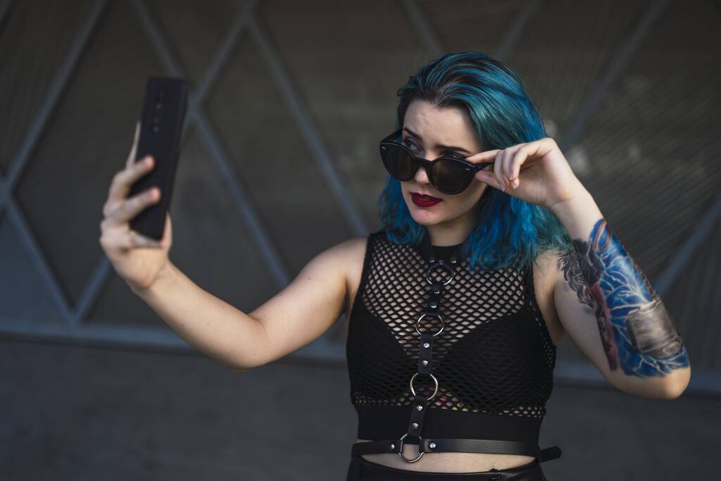 Une femme européenne avec tatouage et cheveux bleus portant des lunettes de soleil et une robe noire sexy prenant un selfie sur le côté d'un bâtiment - Photo, image