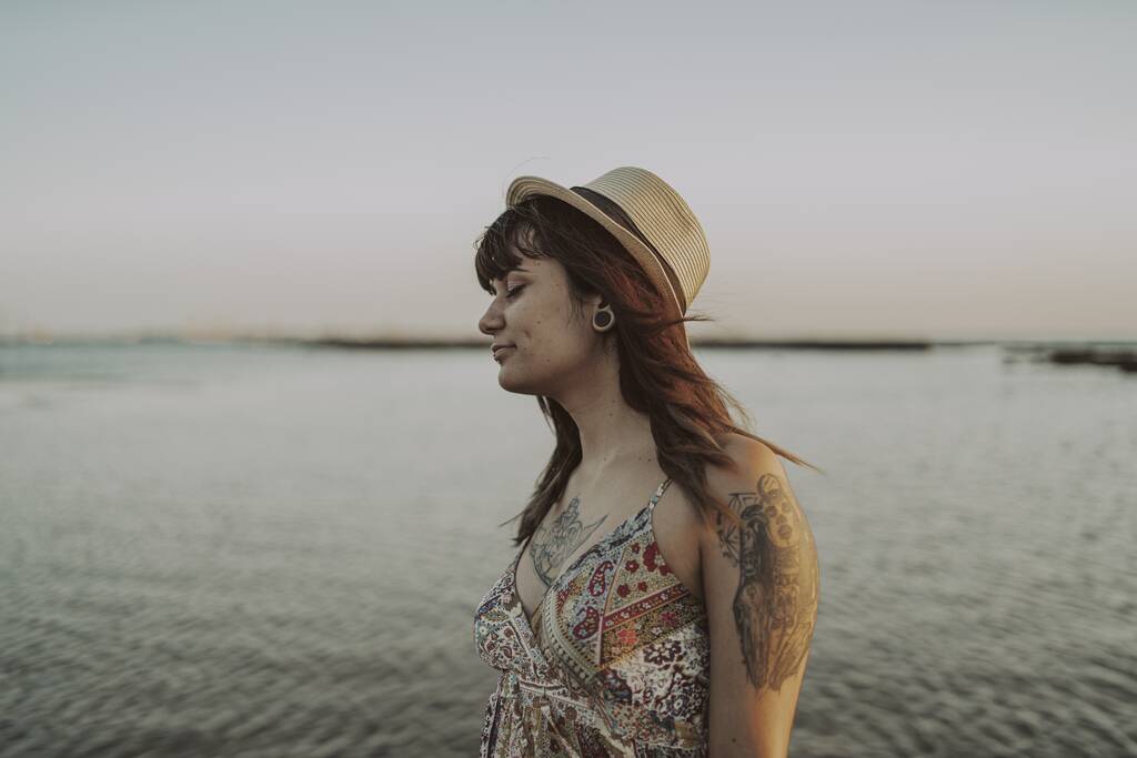 Mladá samice s tetováním v šatech a slaměným kloboukem na rozmazaném mořském pozadí - Fotografie, Obrázek